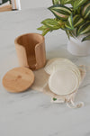 Wooden container with lid, cotton pads in a mesh bag, and a plant on a marble surface